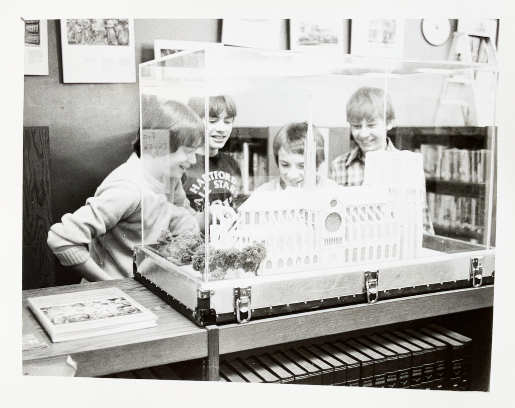 Students gather around a detailed architectural model displayed in a clear case, examining it together in a school library or media center.