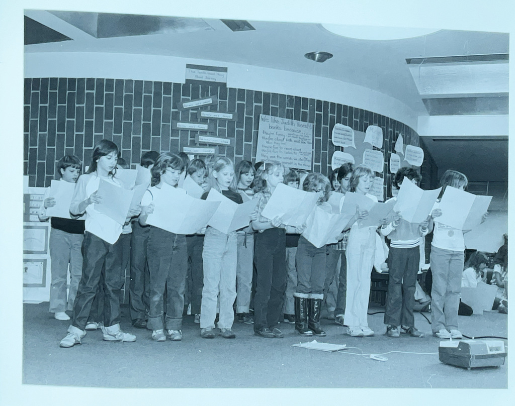 Elementary students stand together indoors holding papers as they participate in a classroom reading or singing activity.