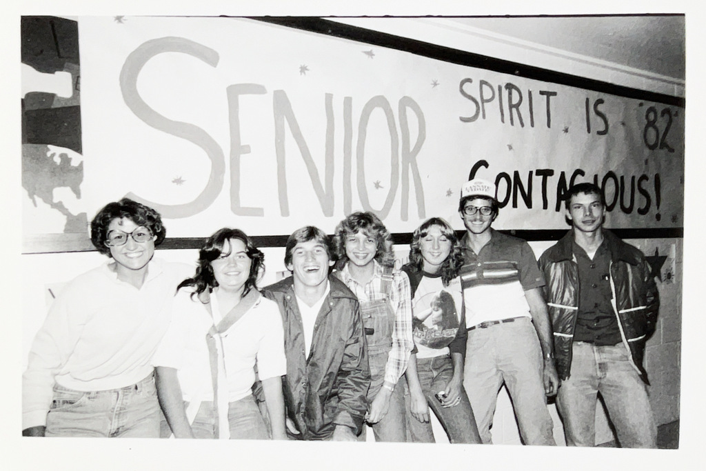 Black-and-white photo of seven students posing together in front of a hand-painted banner reading “Senior Spirit is ’82 Contagious!”