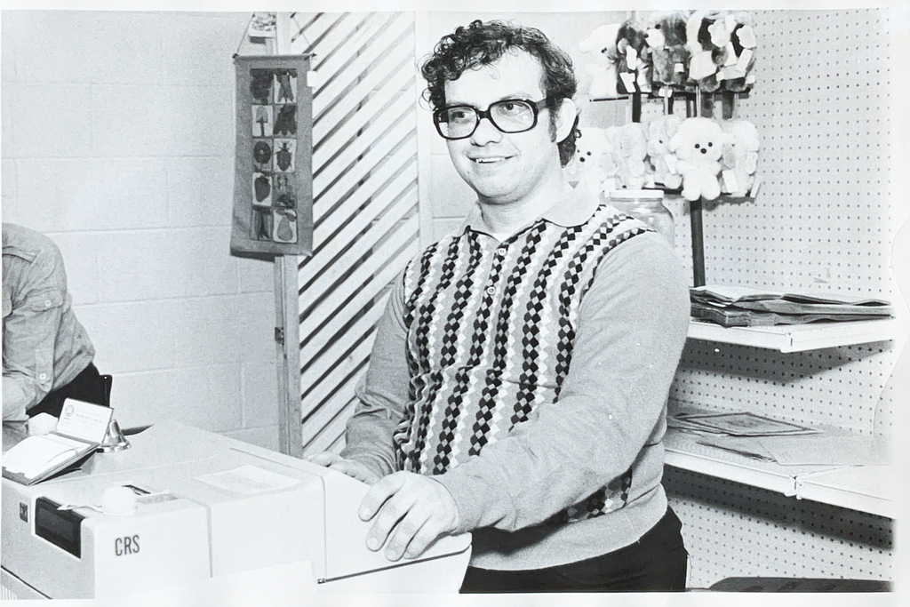 Black-and-white photo of a smiling man wearing glasses and a patterned sweater standing behind a large machine or piece of office equipment.