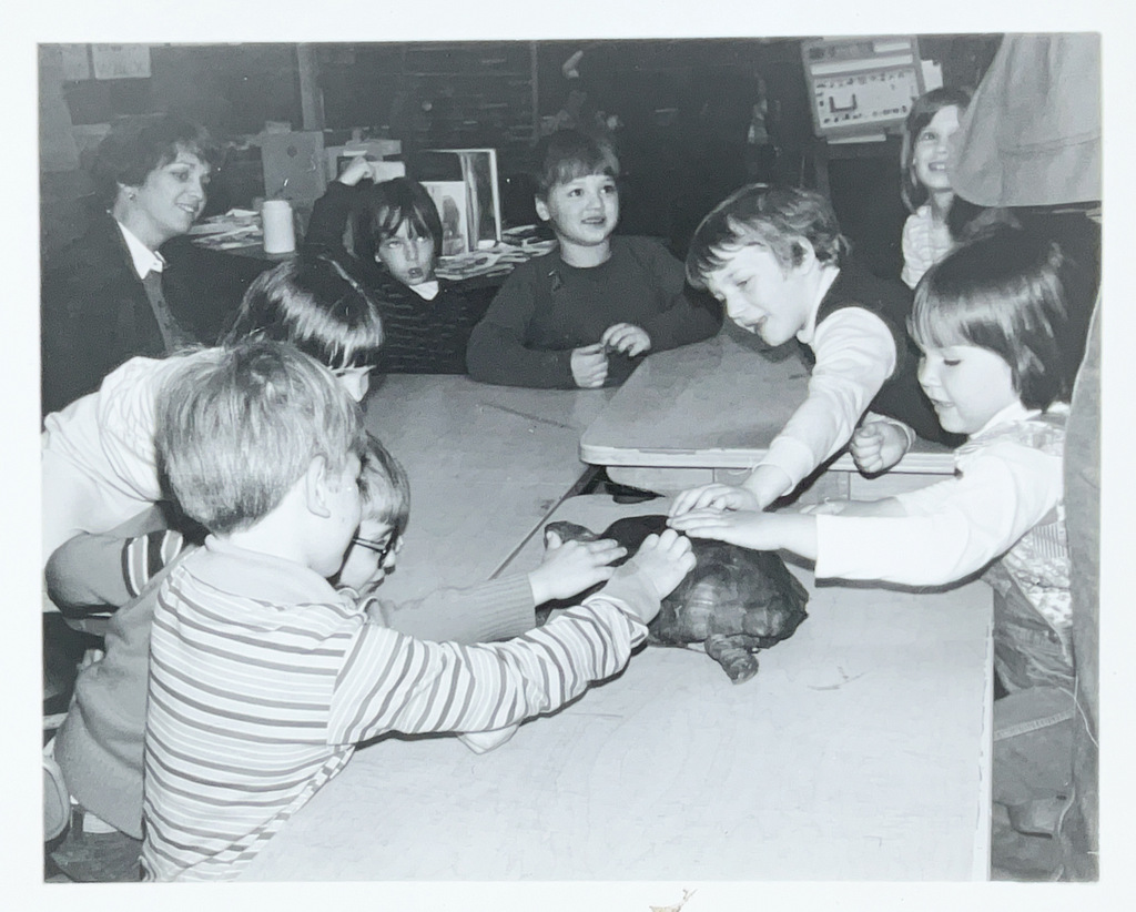 Black-and-white photo of elementary-age children gathered around a classroom table, smiling and reaching toward a turtle while an adult watches from the background.
