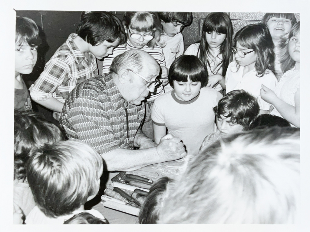 Black-and-white photo of an older man demonstrating an activity at a table while a large group of children gathers closely around to watch.