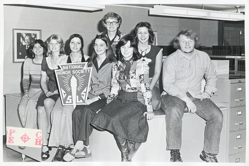 Black-and-white photo of students and an adult posing in a school room with a large National Honor Society emblem displayed in front.
