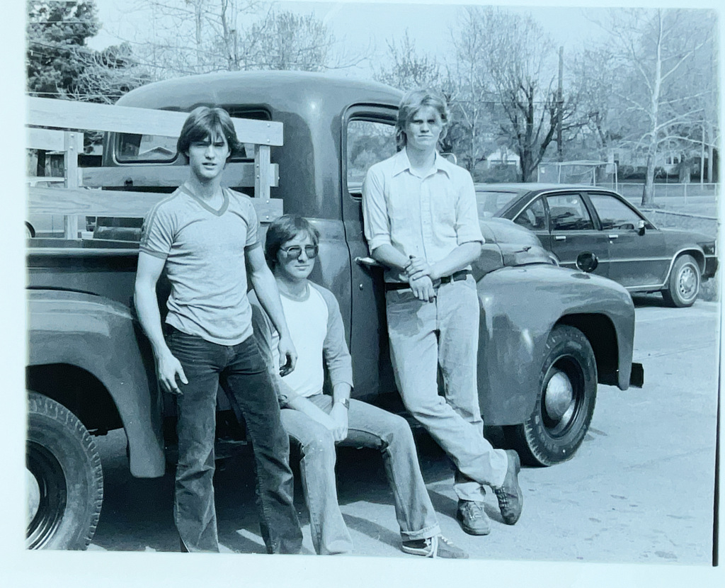 Black-and-white photo of three young men posing beside an older pickup truck in a school parking lot.