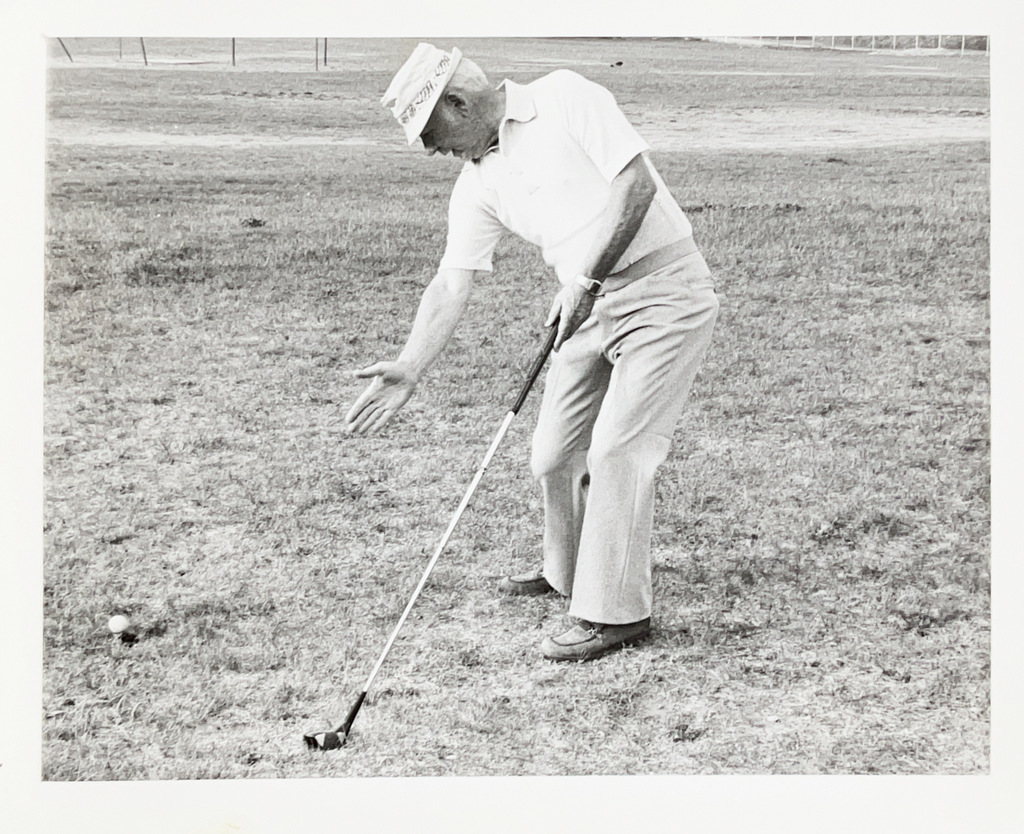 Black-and-white photo of a man on a grassy field preparing to hit a golf ball with a club, extending one hand toward the ball.