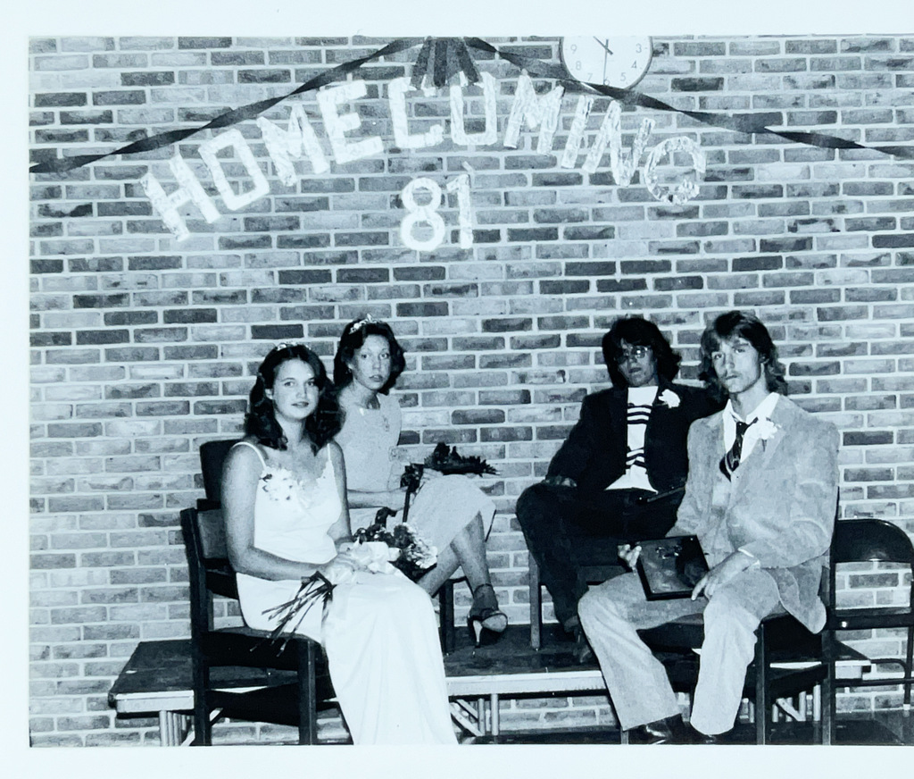 Black-and-white photo of four students seated on a stage beneath decorations reading “Homecoming ’81.” Two students in front wear formal attire and hold flowers.