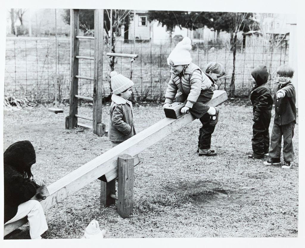 Black-and-white photo of children bundled in coats and hats playing outdoors around a wooden seesaw on a school playground.