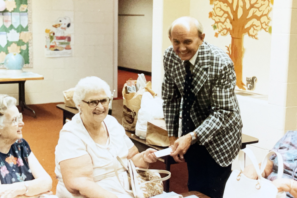 An older man in a checkered suit smiles while handing a small card or paper to an older woman seated at a table during a school gathering. Other attendees and bags are visible around them.