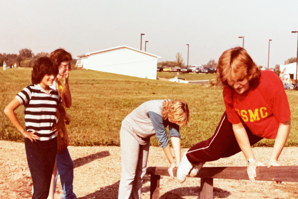 Students participate in an outdoor obstacle-course activity, with one student climbing over a wooden barrier while others wait nearby on the school grounds.