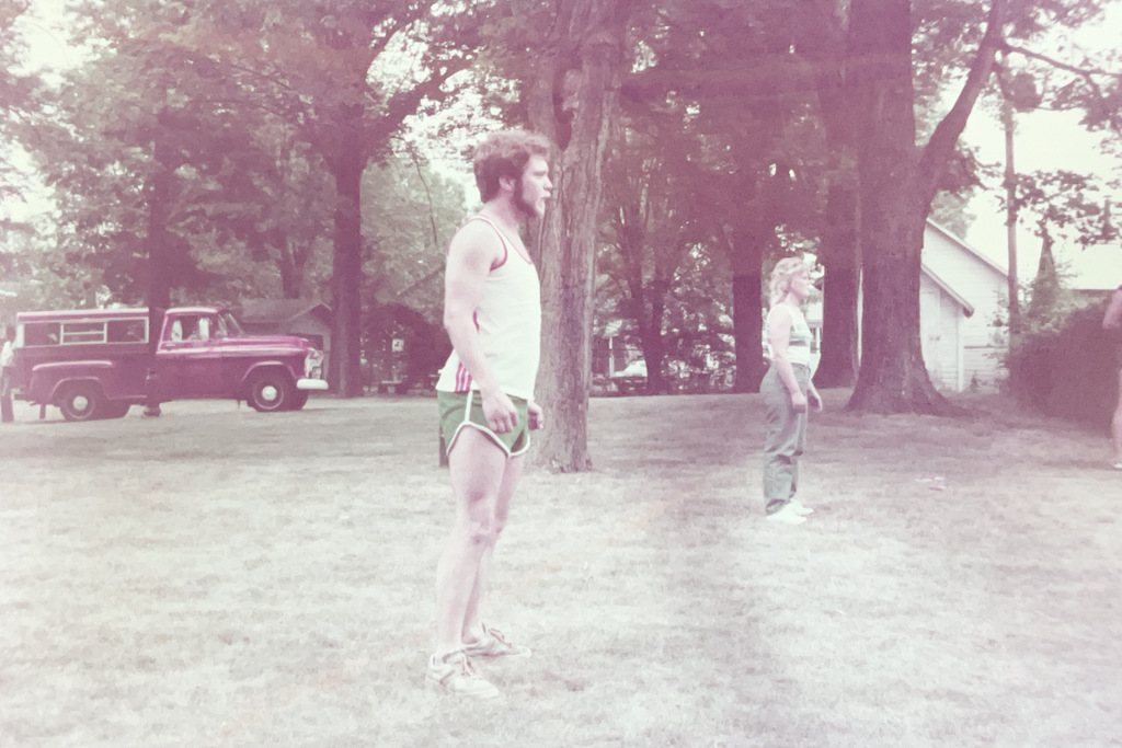 A man wearing athletic clothing stands outdoors on a grassy field near another person, with trees, a house, and a parked pickup truck in the background.