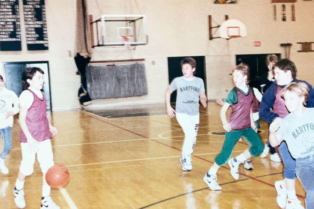 Students play basketball in a school gymnasium, with several players wearing practice pinnies and Hartford basketball shirts.
