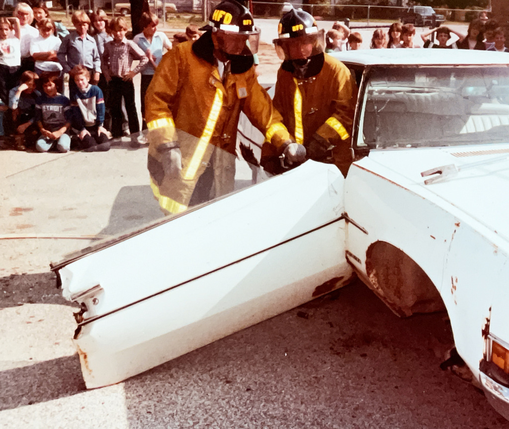 Two Hartford firefighters in protective gear work beside a heavily damaged white car as a group of students and adults watch from behind.
