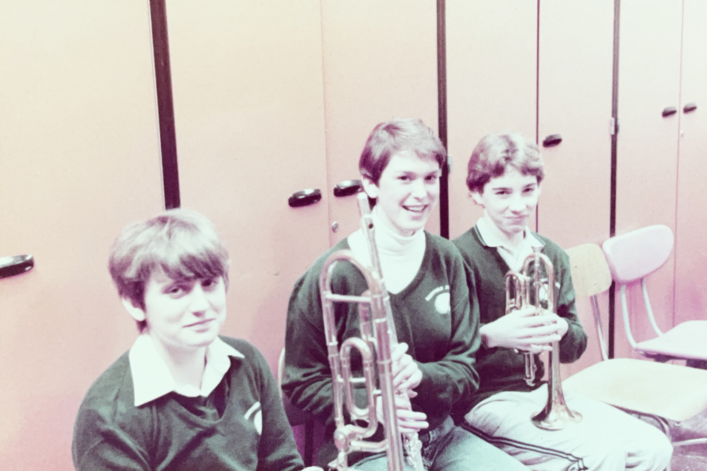 Three students sit together holding brass instruments, including trombones, in what appears to be a school band room.