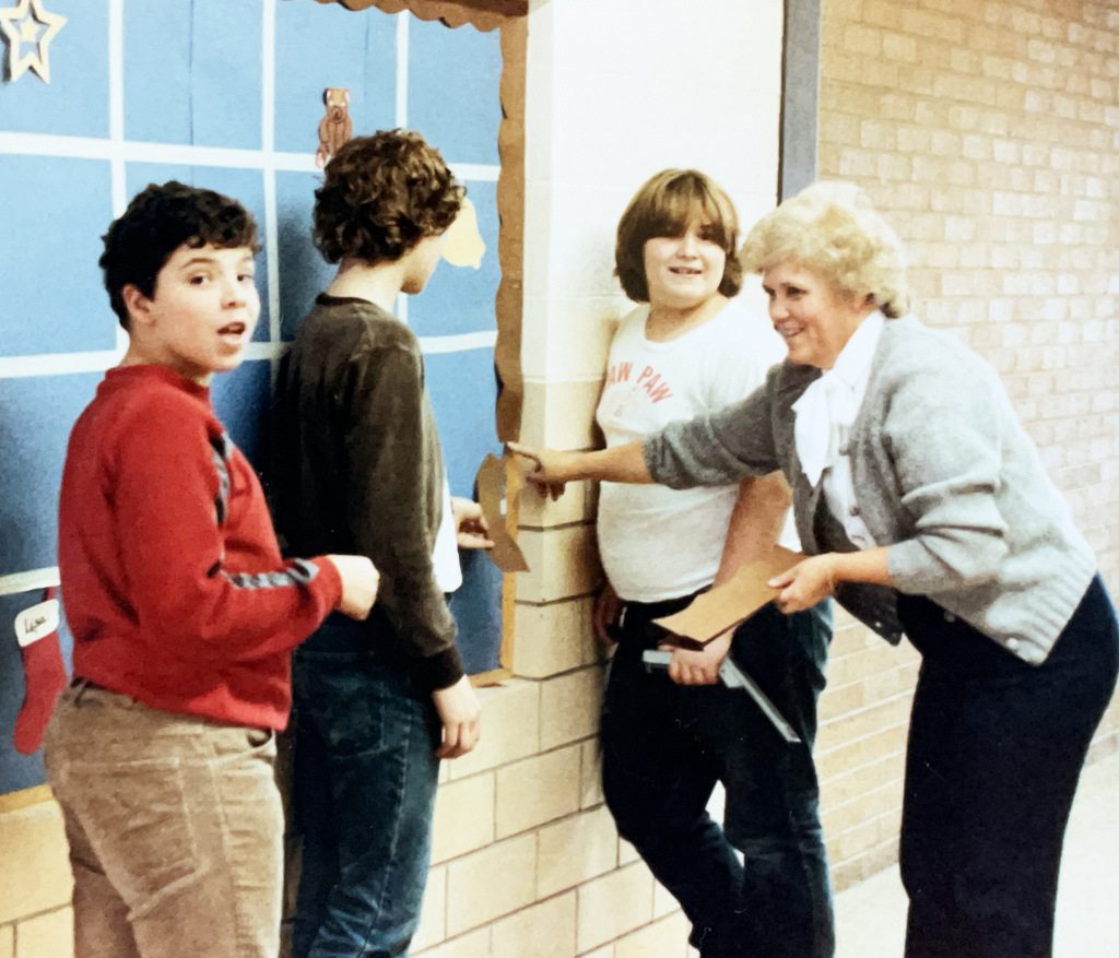 A staff member interacts with three students beside a decorated school bulletin board. The group appears to be participating in a classroom or school activity.