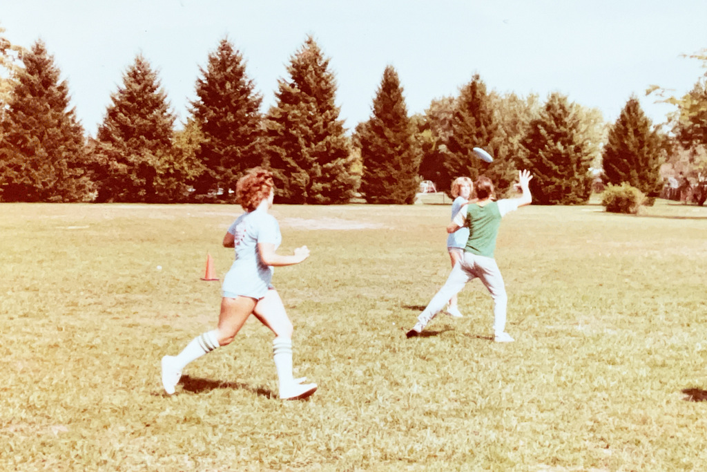 Students play a game outdoors on a grassy field, with one student throwing a flying disc while others run nearby.