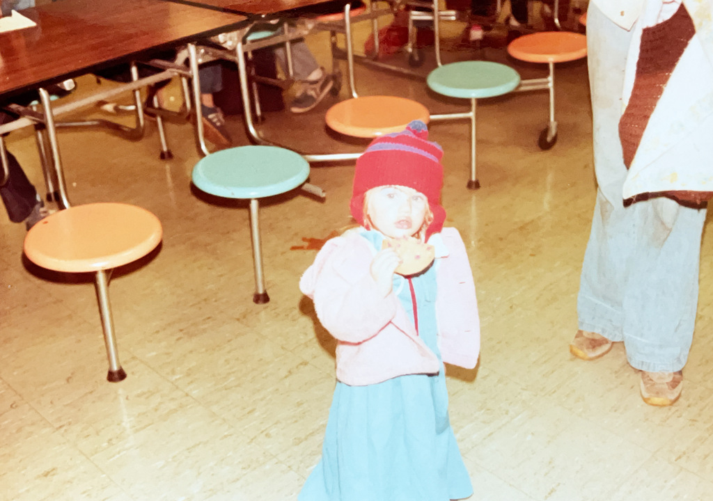 A young child wearing a winter hat, coat, and dress stands in a school cafeteria while eating a cookie. Colorful round cafeteria stools and tables are visible in the background.