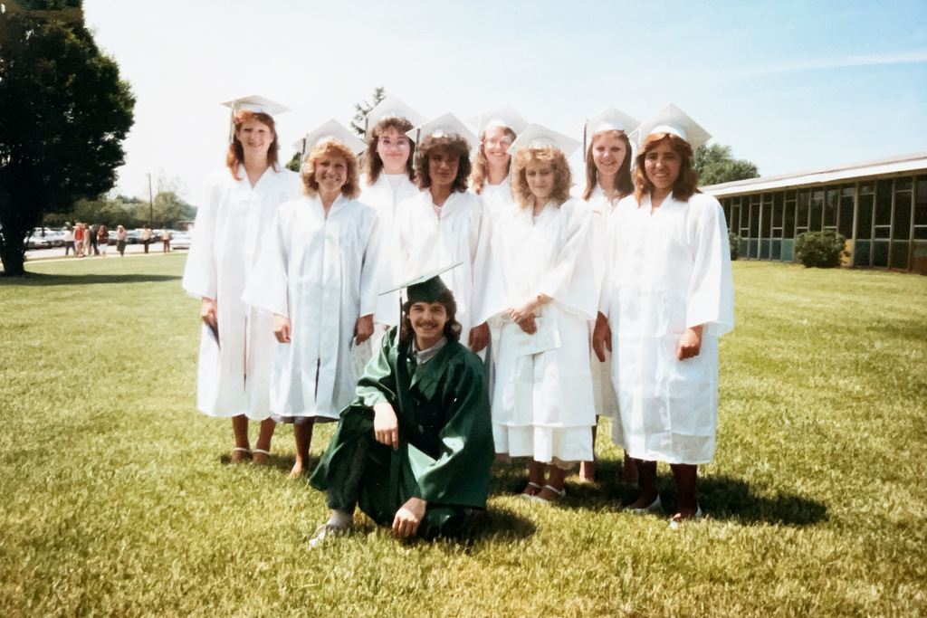 A group of graduates pose together outdoors on the school grounds in caps and gowns. Most wear white graduation gowns, while one graduate in a green cap and gown kneels in front.