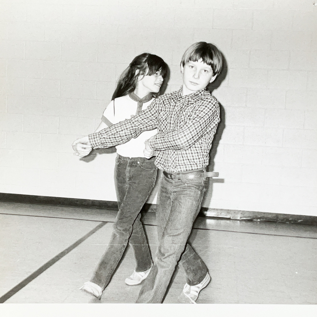 Black-and-white photo of two students standing in a school gym. A boy in a plaid shirt poses in front while a girl stands behind him, appearing to participate in a dance or movement activity.