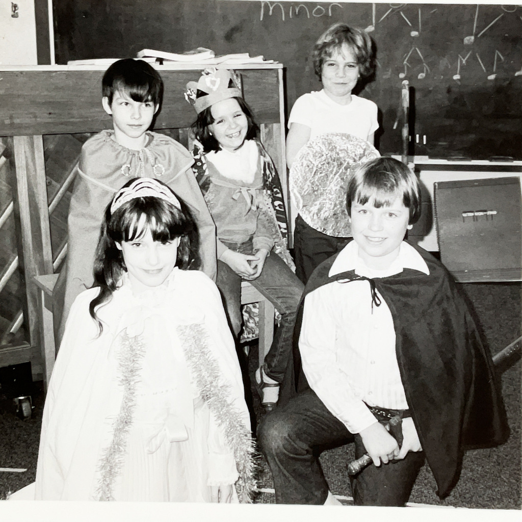 Black-and-white photo of five students dressed in costumes for a classroom performance. Students wear capes, crowns, and other handmade costume pieces, with musical notes drawn on the chalkboard behind them.