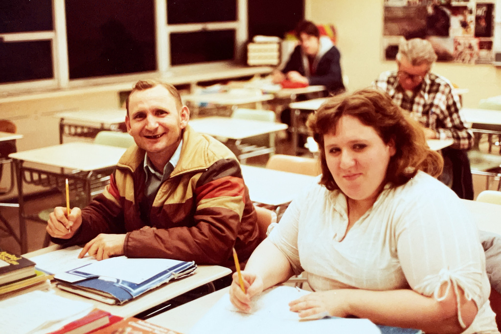 Color photo of two adults seated at desks in a classroom, smiling toward the camera while holding pencils. Other adults are seated at desks in the background.