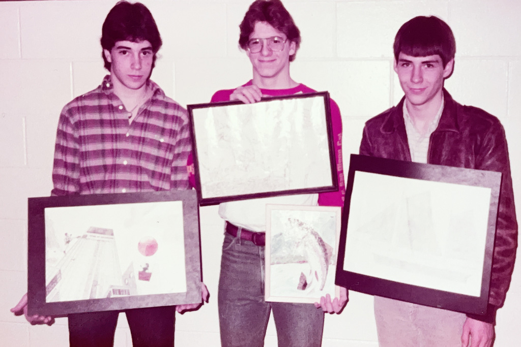 Vintage color photo of three students standing against a school wall and displaying framed pieces of artwork.