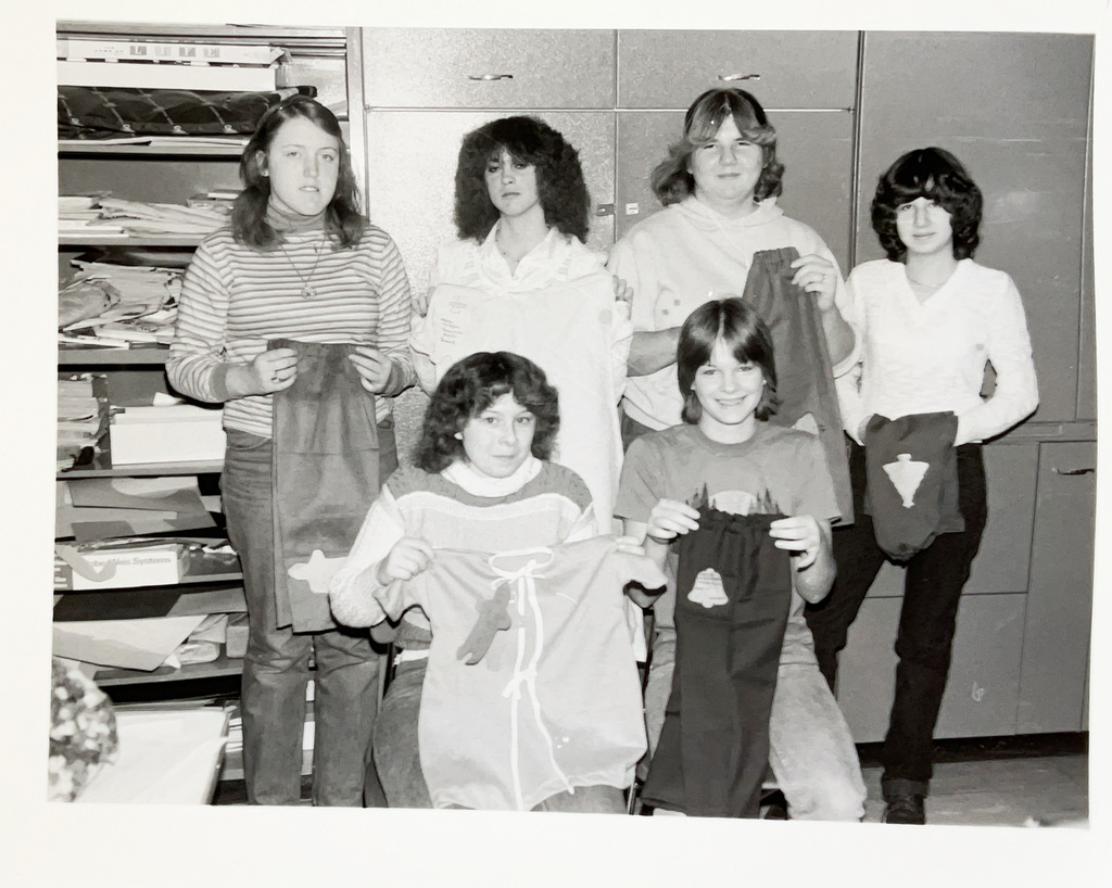 Black-and-white photo of six students posing in a classroom while displaying handmade fabric projects. Four students stand in the back while two sit in front holding their creations.