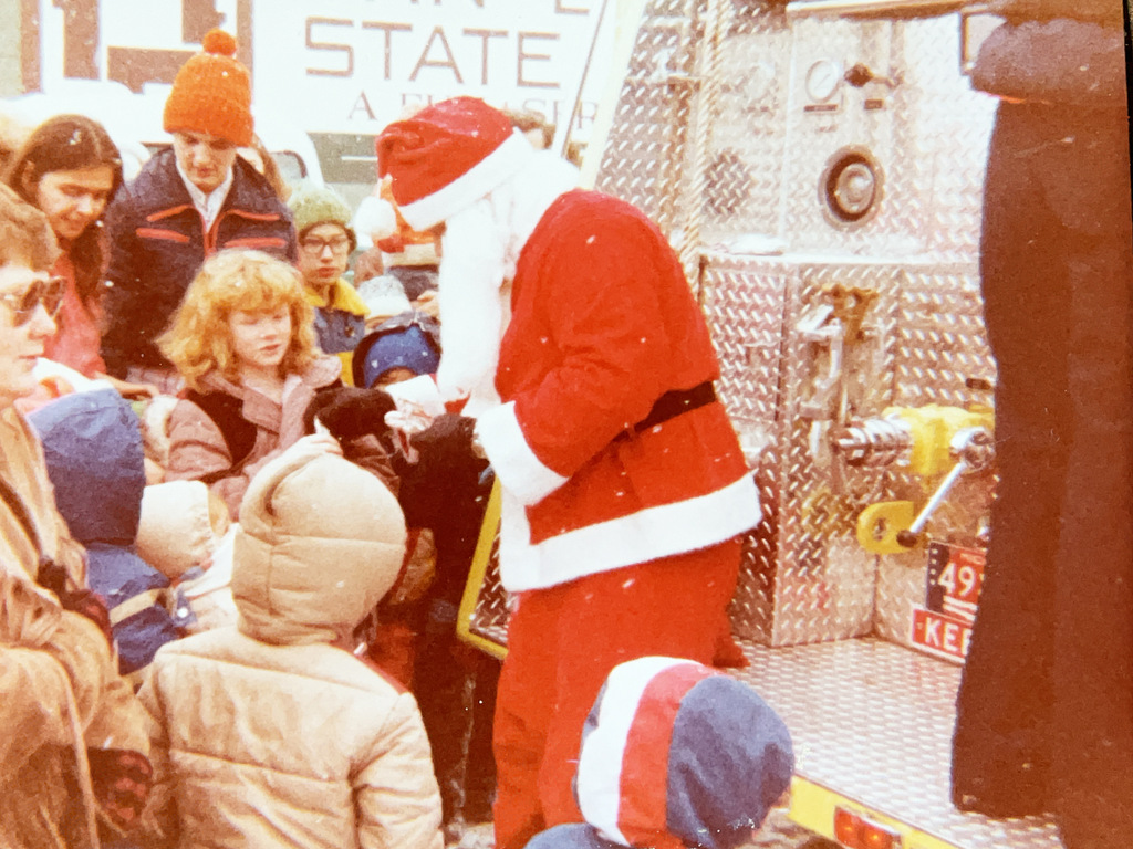 Vintage color photo of children gathered around Santa Claus beside a fire truck during a winter community event. Santa appears to be handing something to a child as families gather nearby.