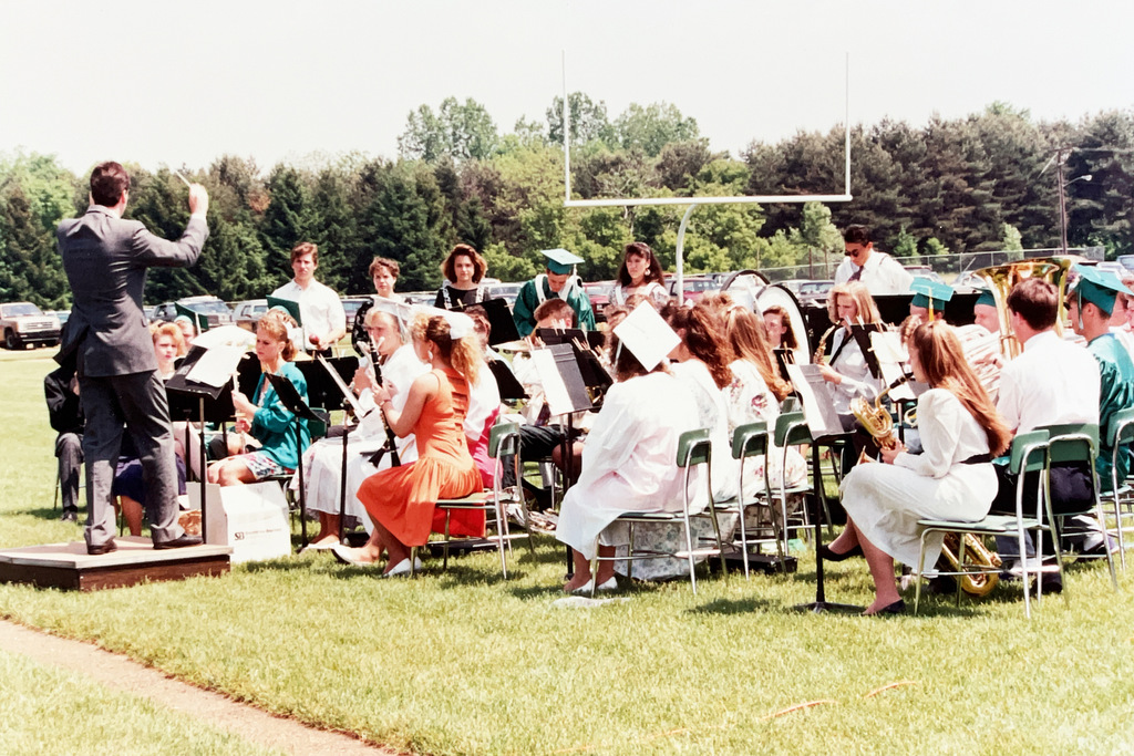 Color photo of a school band performing outdoors on a football field during a graduation ceremony. A conductor leads the musicians while graduates in caps and gowns sit among the ensemble.