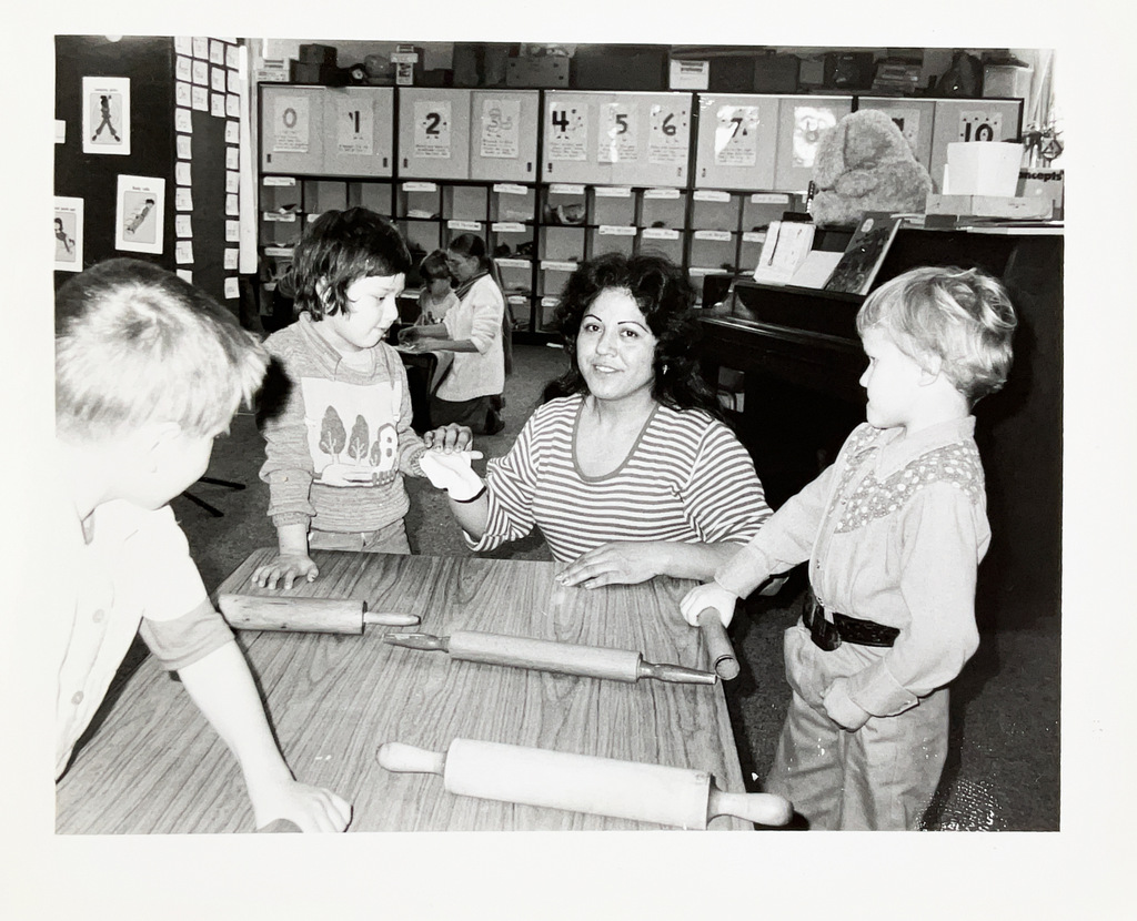 Black-and-white photo of a teacher working with young students around a classroom table. Several rolling pins are on the table as the group participates in a hands-on activity.