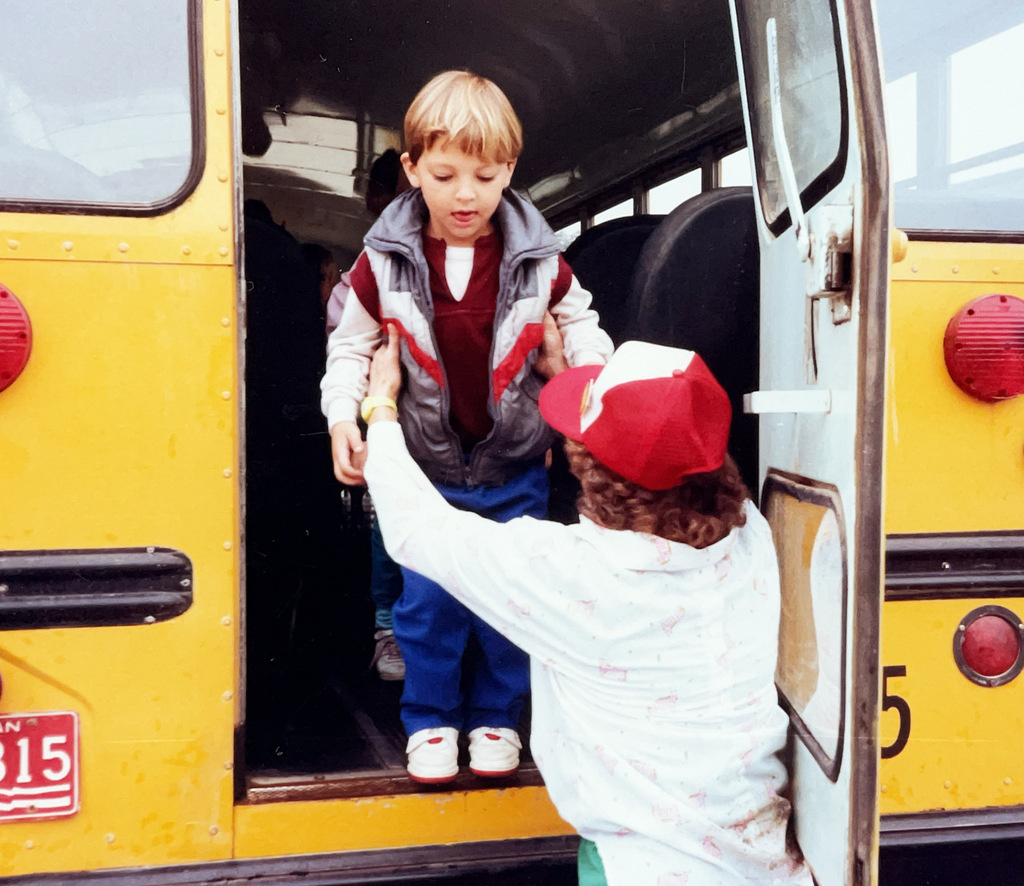 Color photo of a young student stepping off a yellow school bus while an adult reaches up to help him down the steps.