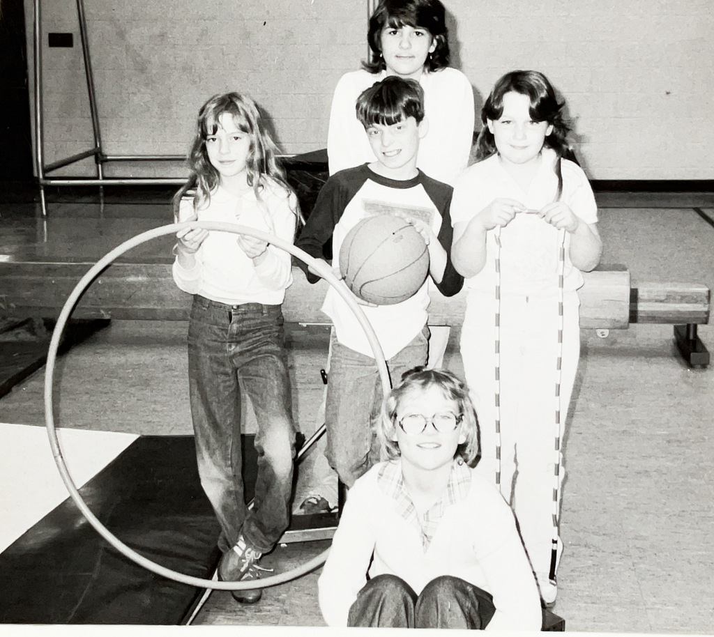 Black-and-white photo of five students posing in a school gym with physical education equipment. One student holds a basketball, another holds a large hula hoop, and another holds a jump rope.