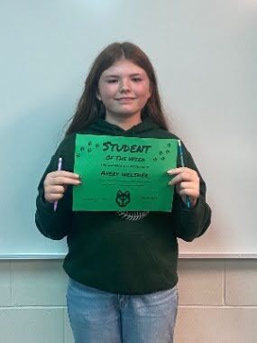 Student stands in front of a classroom wall holding a green “Student of the Week” certificate.