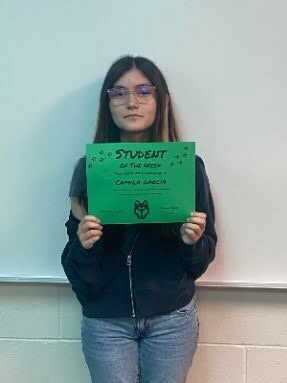 Student stands in front of a classroom wall holding a green “Student of the Week” certificate.