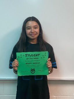 Student stands in front of a classroom wall holding a green “Student of the Week” certificate.