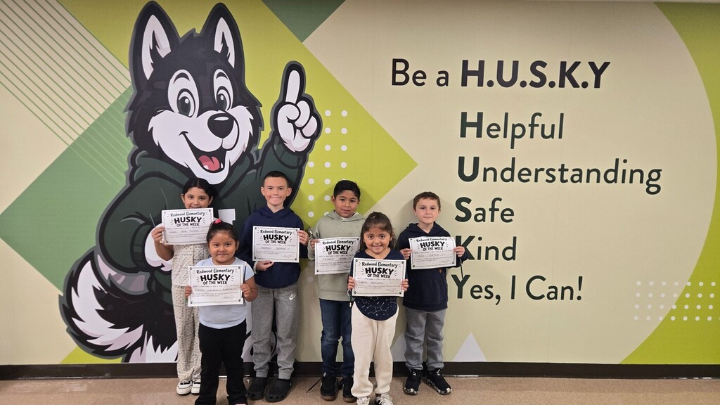 Six Redwood Elementary students pose together with their “Husky of the Week” certificates in front of a colorful school mural featuring the Husky mascot and the H.U.S.K.Y. values: Helpful, Understanding, Safe, Kind, and “Yes, I Can!”