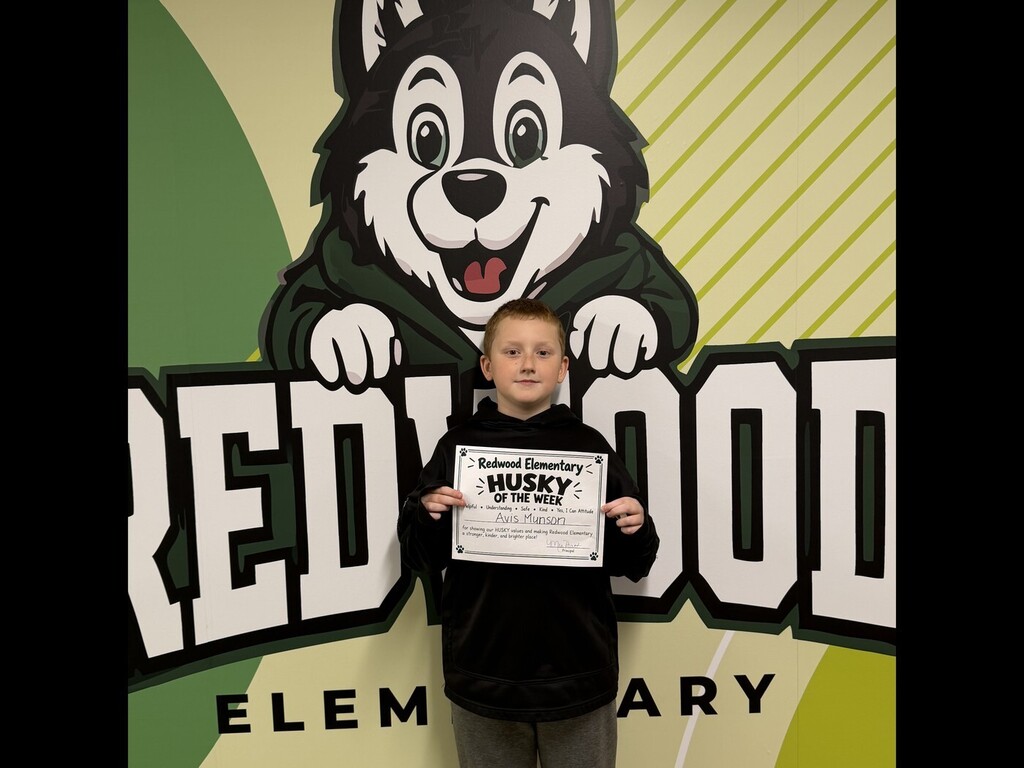 A Redwood Elementary student wearing a black hoodie stands in front of the school’s Husky mural while displaying his “Husky of the Week” certificate.