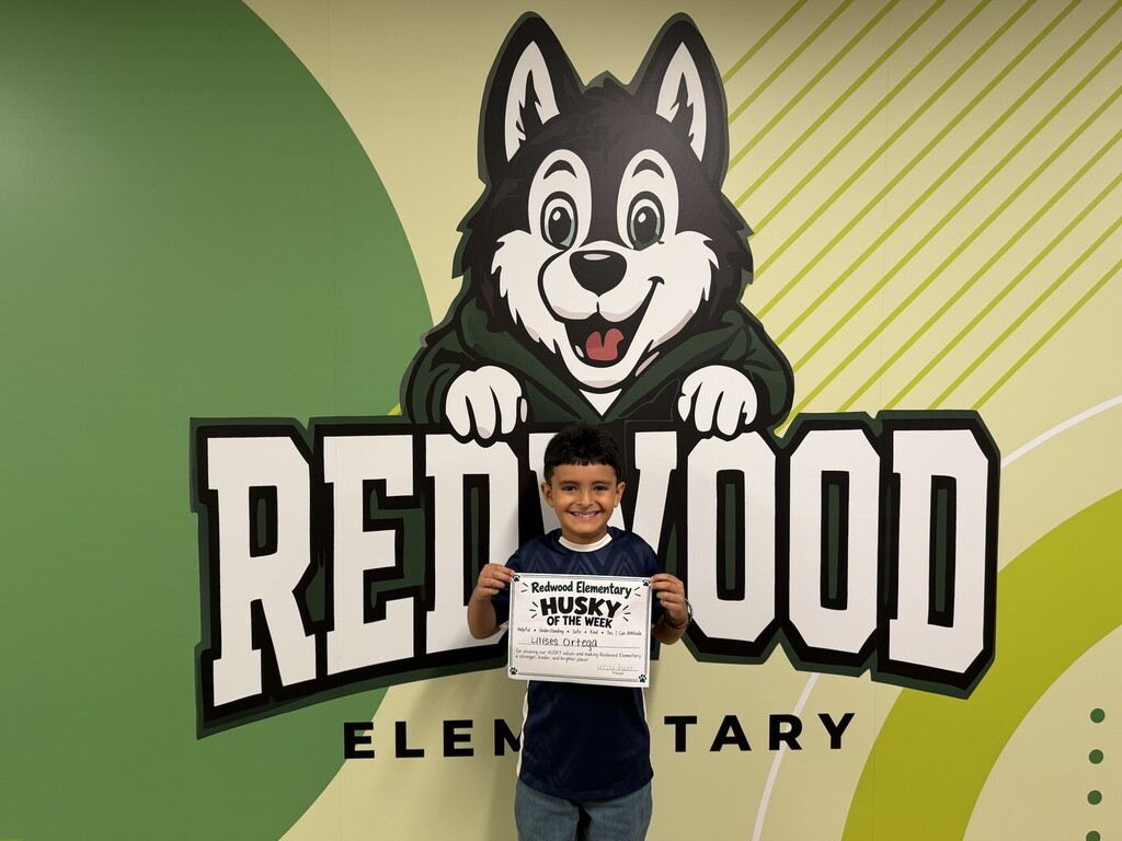 A smiling Redwood Elementary student stands in front of the school’s Husky mural while holding a “Husky of the Week” certificate.