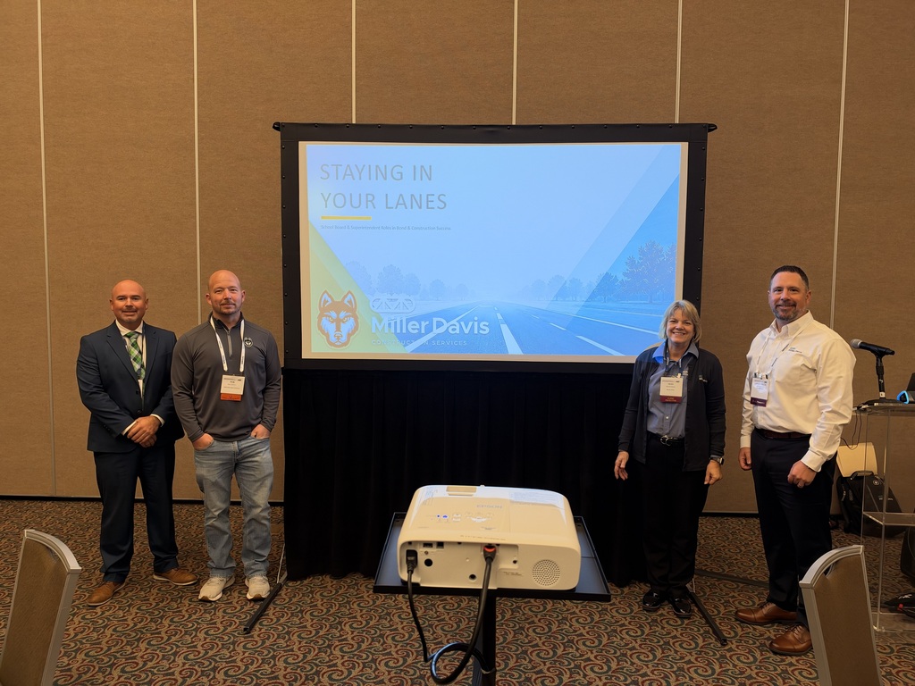 Hartford Public Schools Board of Education member Rob Sheffey and Superintendent Nick Blackmer pose with two representatives from Miller Davis Construction Services during the Michigan Association of School Boards Leadership Conference. The four presenters stand on either side of a large projection screen displaying a presentation titled “Staying in Your Lanes,” featuring the Hartford Huskies logo and Miller Davis branding. The presentation focused on the school bond process, from initial vision through construction, and the roles and responsibilities of school board members and the community. The group stands in a conference room with wood-paneled walls, patterned carpeting, and a projector positioned in front of the screen.
