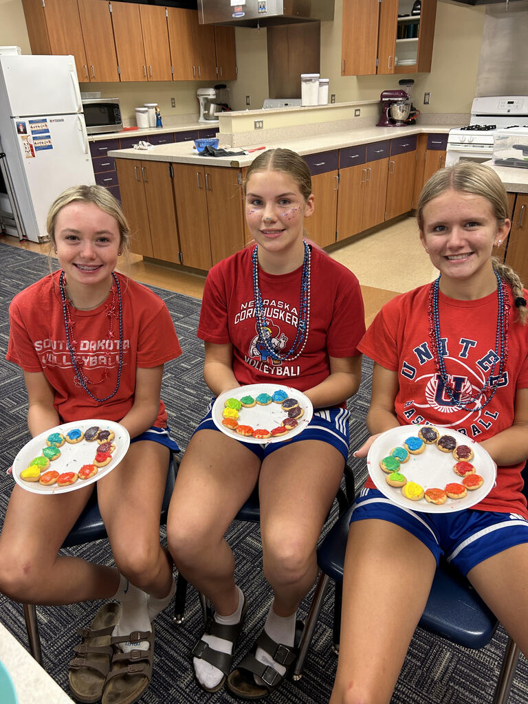 Three smiling students in red team shirts hold colorful decorated plates in a kitchen, celebrating creativity and team spirit.