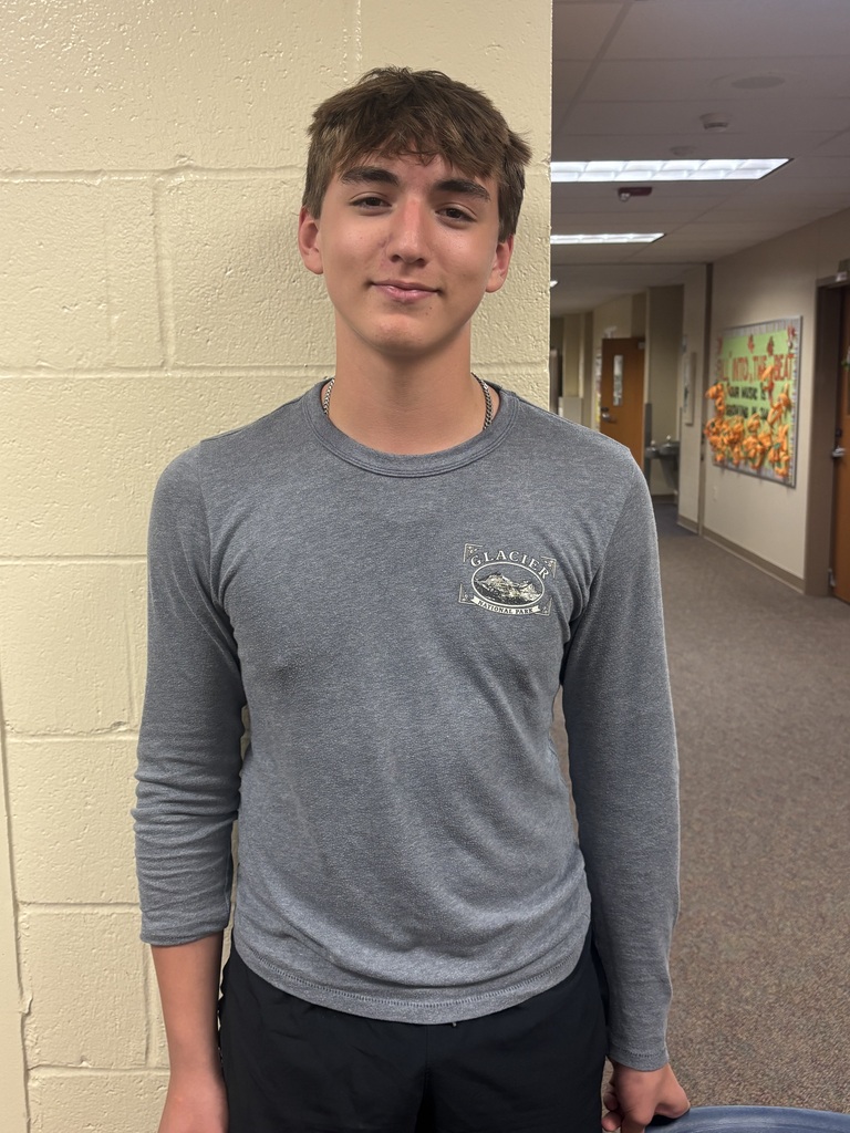 Teenage boy in a gray long-sleeve shirt and black pants stands against a school hallway wall, giving a calm, slight smile in a casual portrait.