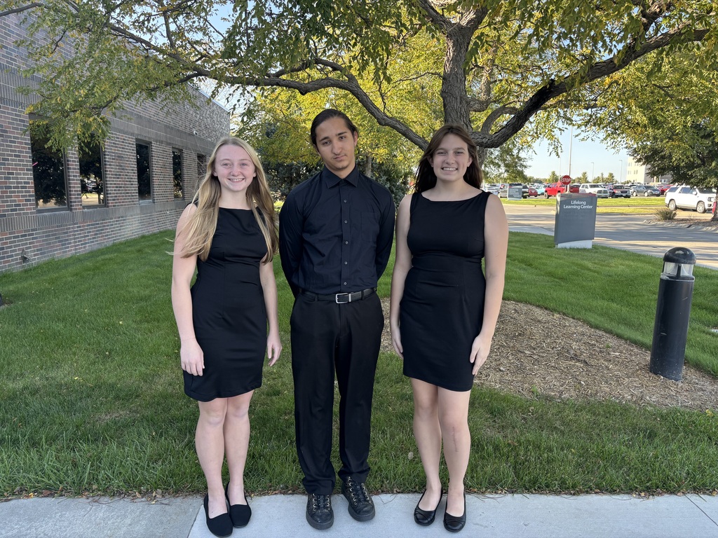 Three young adults in black formal outfits stand together beneath a leafy tree outside an office building, smiling in a calm, sunny setting.