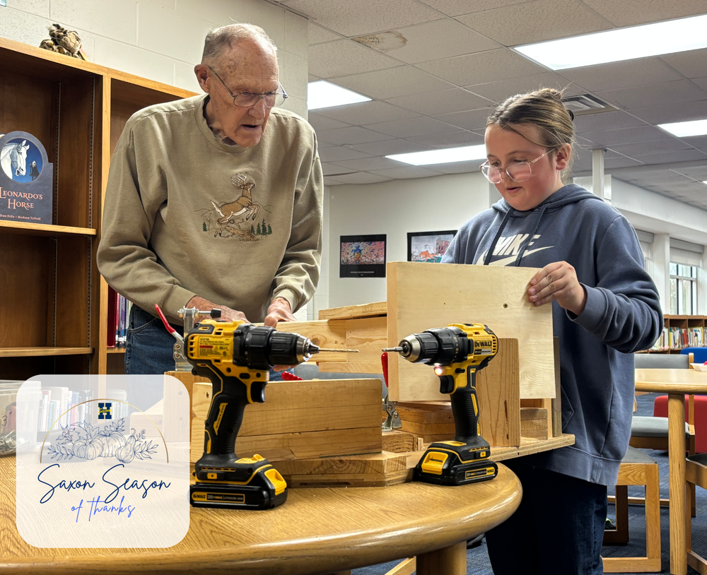 A man stands next to a young female student. They are arranging pieces of wood to build a birdhouse