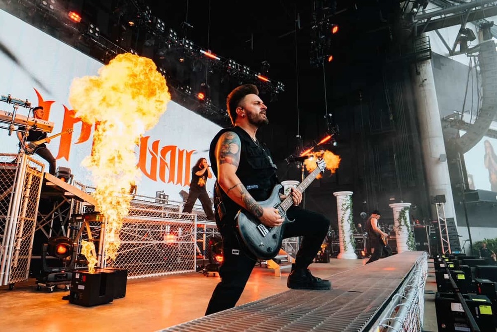 A guitarist stands on stage playing as flames rise up from behind him.