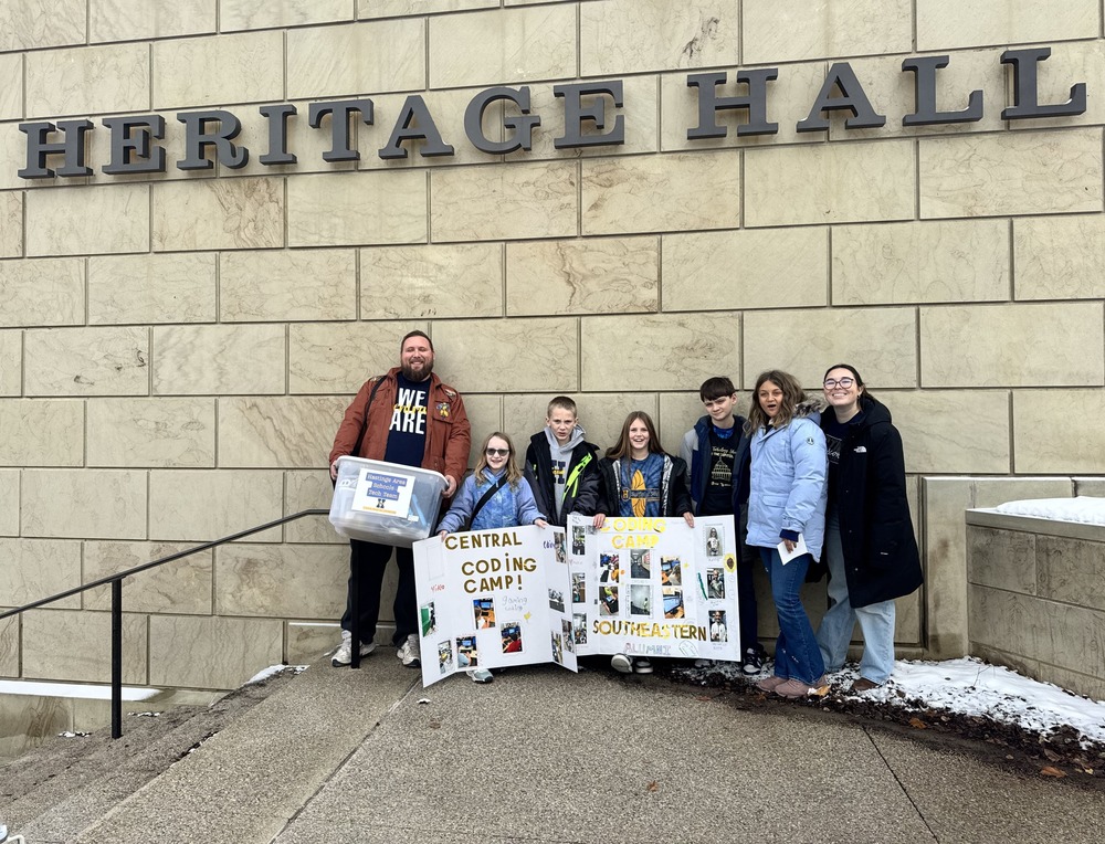 A group of students and teachers stand in front of a sign that says Heritage Hall. They are outside and bundled up in warm clothing