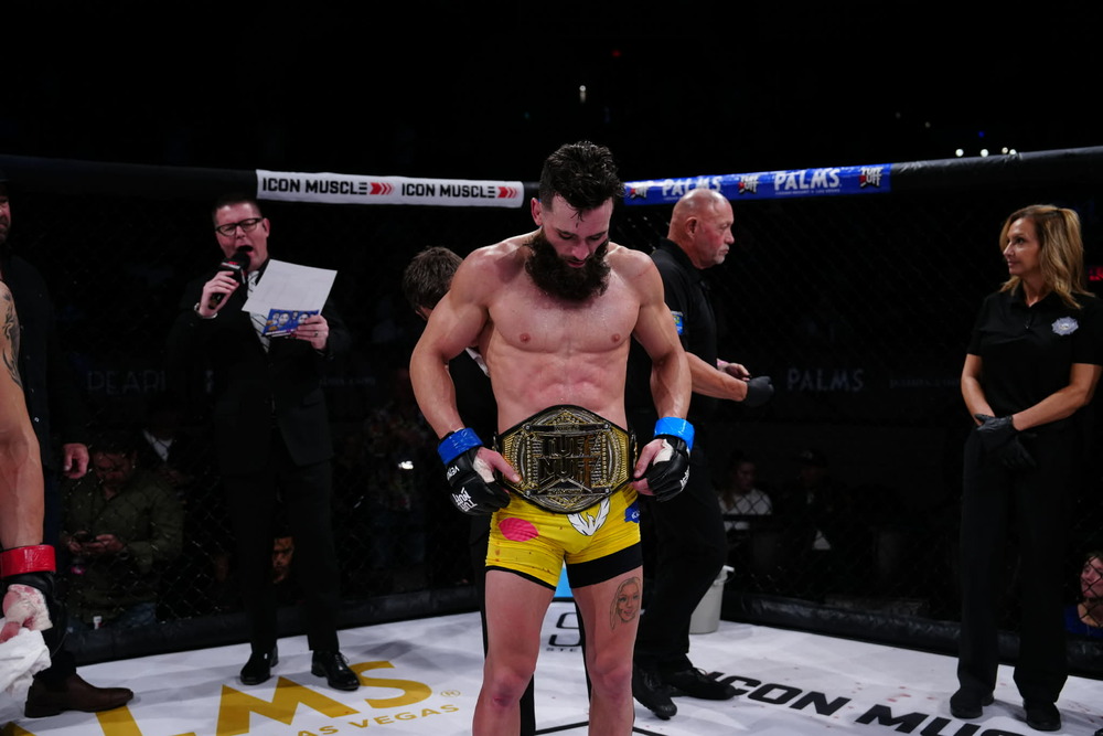 A shirtless MMA fighter stands in the center of the ring with a belt around his waist.