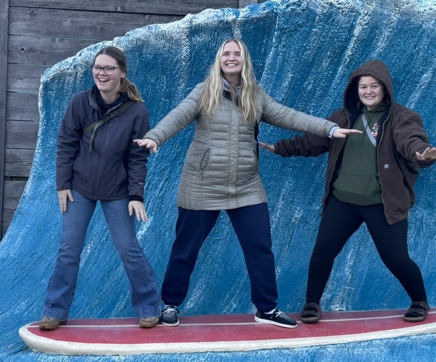 Students posing on a surf board on top of what appears to be fake water. They're smiling big.