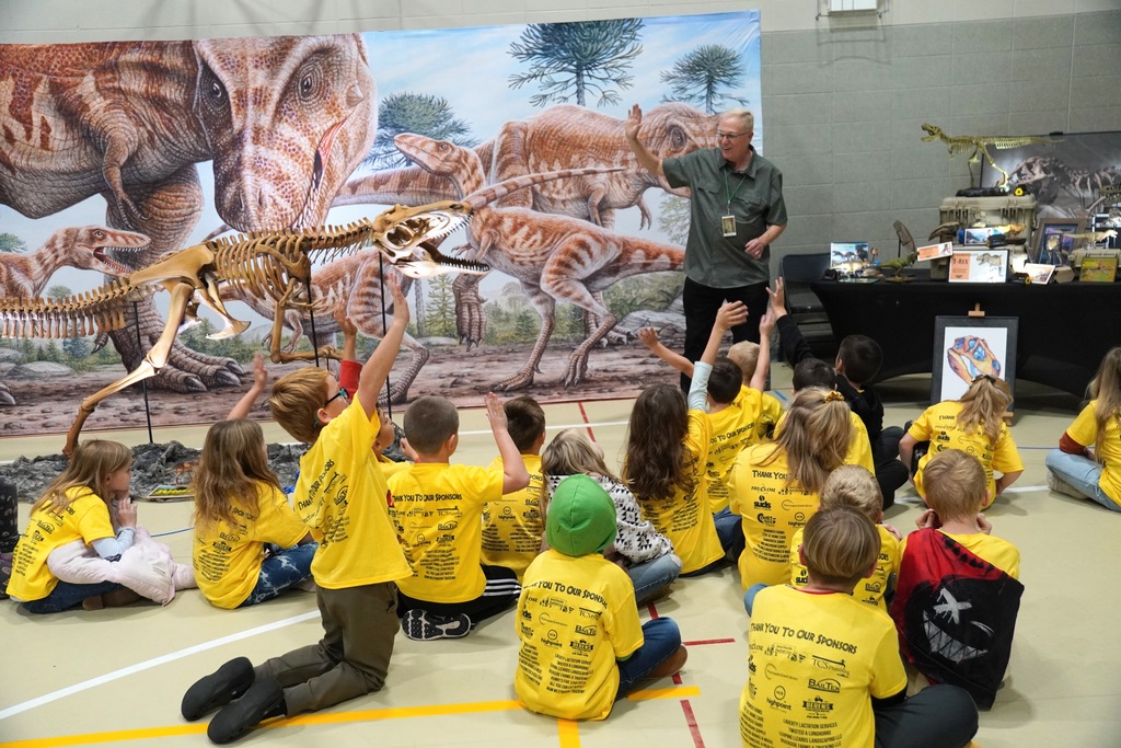 A  man presents dinosaur fossils and learning materials to a group of kids. Some kids are raising their hands.