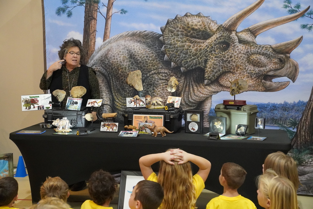 A woman is standing behind a table of dinosaur learning materials and she is speaking to kids.