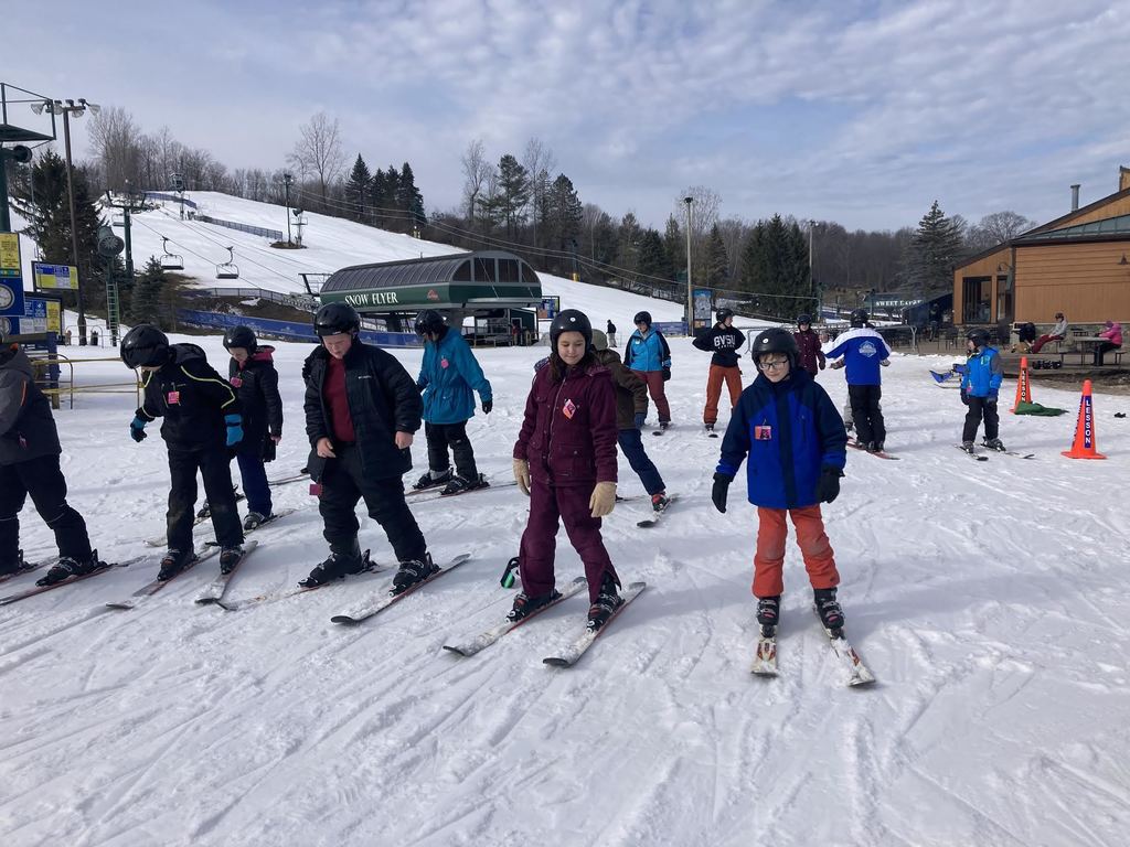 Kids standing shoulder to shoulder, bundled up in winter gear while standing on snow skis. They also have helmets on.