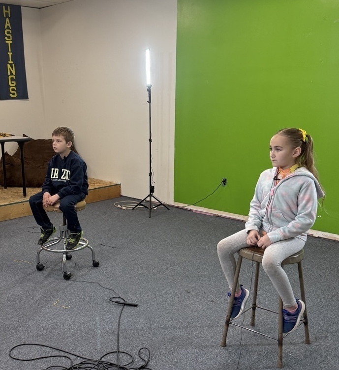 Two young students sit in front of a green screen looking ahead.